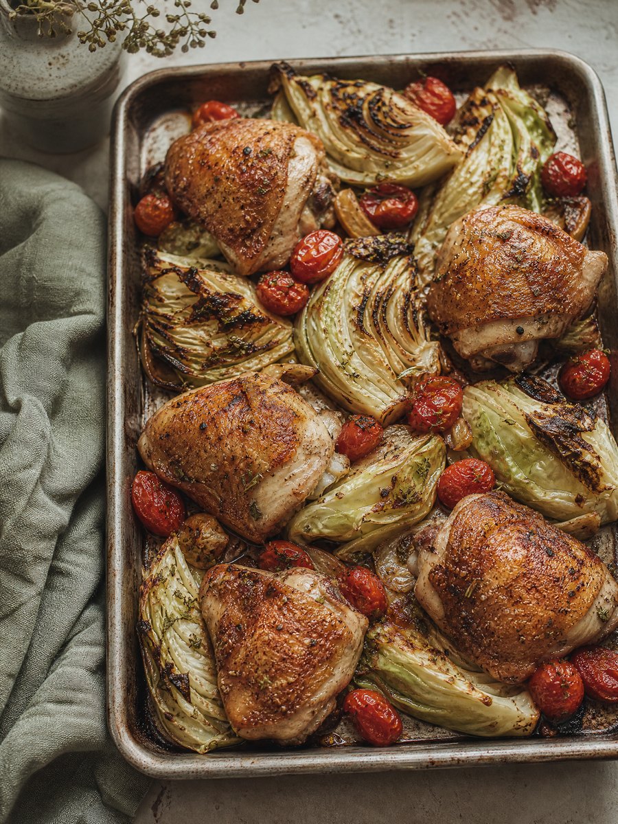 A rimmed metal baking sheet holding roasted bone-in chicken thighs with crispy skin, surrounded by charred wedges of green cabbage.
