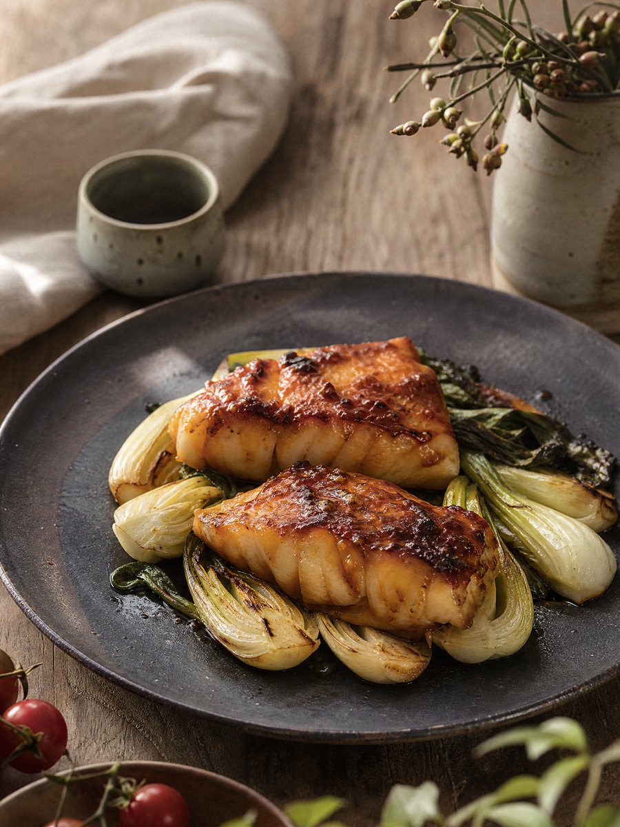 Two fillets of miso-glazed cod resting on a bed of roasted baby bok choy on a dark ceramic plate, set on a wooden table.